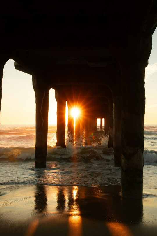 The sun is setting under a pier at the beach